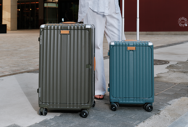 Two suitcases, one gray and one teal, on wheels with a person in light clothing in the background.